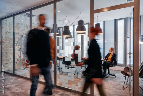 In the dynamic hustle and bustle of a business environment, a group of young business professionals walk down the corridor next to their office, while their colleagues collaborate inside, symbolizing