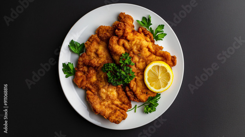Overhead shot of breaded and fried wiener schnitzel with parsley and a lemon slice on a white plate against a black background