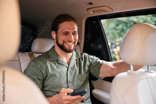 Smiling man using smartphone in a car interior with earbuds, casual outfit, relaxed atmosphere