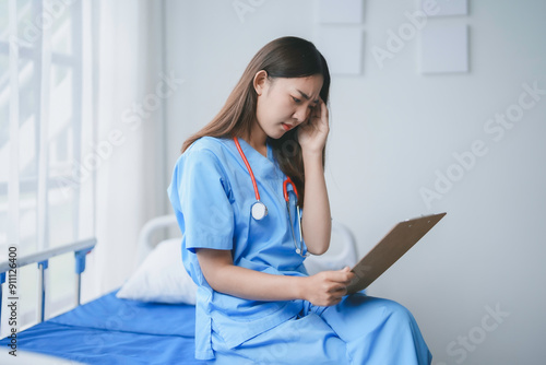 Young doctor in scrubs is sitting on a hospital bed, holding a medical chart and experiencing a headache