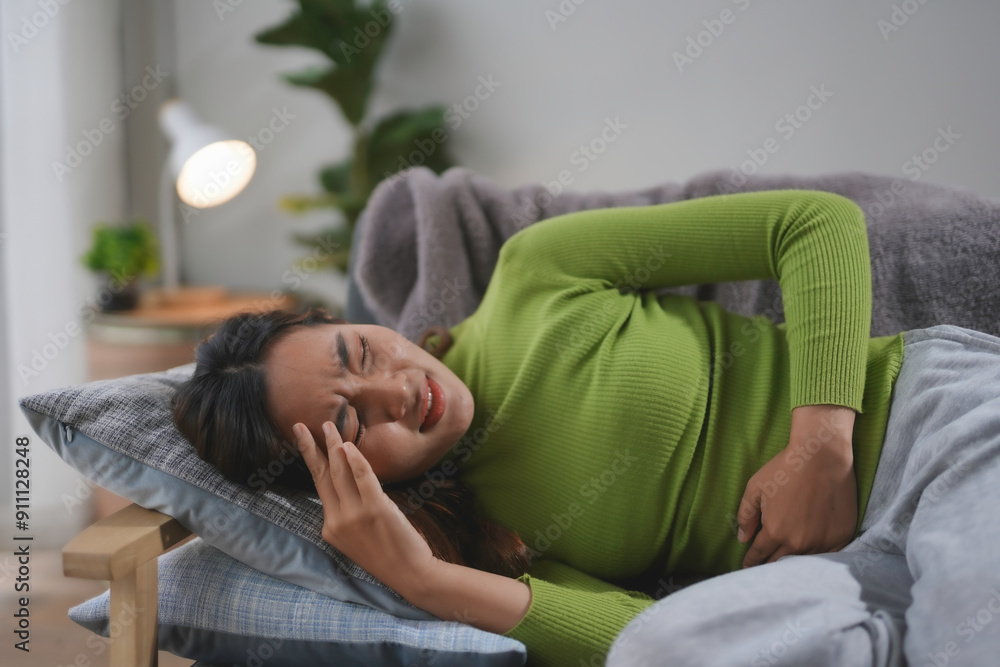 Young woman is experiencing intense stomach pain while lying on her side on the sofa, her hand resting on her abdomen Young woman is experiencing intense stomach pain while lying on her side on the sofa, her hand resting on her abdomen