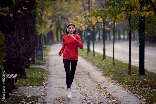 Young beautiful woman running in autumn park and listening to music with headphones on smartphone