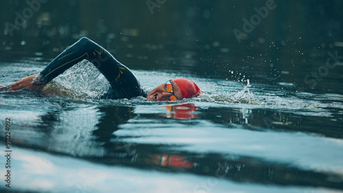Bild auf Leinwand Focused swimmer in wetsuit and red cap powers through the water with orange buoy in tranquil river