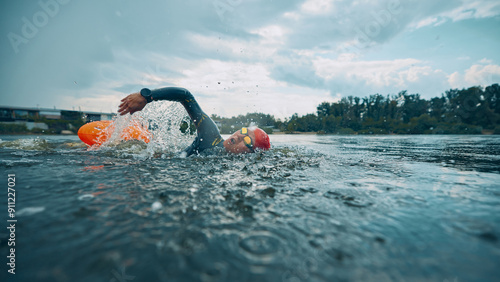 Konstfotografi Gliding through river water, female athlete in black wetsuit and red swim cap accompanied by orange buoy