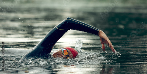 Bild auf Leinwand Triathlete in red swim cap and black wetsuit practices freestyle stroke, arms cutting through water, creating ripples on river's surface
