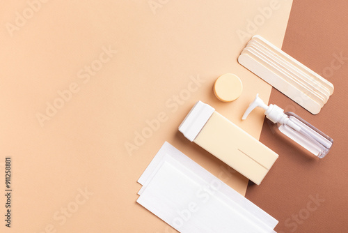 Creative composition with hair removal tools arranged on beige-brown background with copy space. Wax strips, body lotion, honey soft wax cartridge, wooden spatulas for smooth and silky skin.