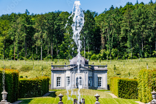 Water jet gushing out in foreground from fountain in Freyr Castle gardens, Renaissance style building in background, lush trees against blue sky, sunny summer day in Namur province, Wallonia, Belgium photo