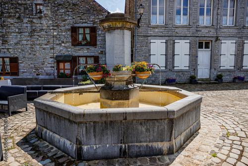 Fototapeta Naklejka Na Ścianę i Meble -  Old fountain in public square in picturesque village of Hierges, flower pot holders with flower pots, buildings with stone walls in background, sunny summer day in the Ardennes departement of France