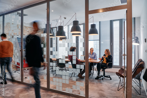 In the dynamic hustle and bustle of a business environment, a group of young business professionals walk down the corridor next to their office, while their colleagues collaborate inside, symbolizing