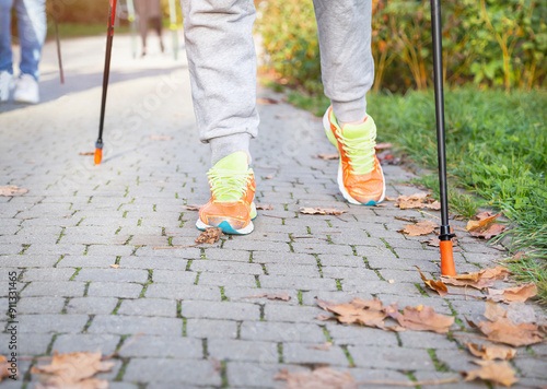 Legs of group of people in sport wear and with walking sticks for popular nordic walking in the autumnal park. Scandinavian trekking sticks hilking, healthy lifestyle. Sneakers shoes. photo