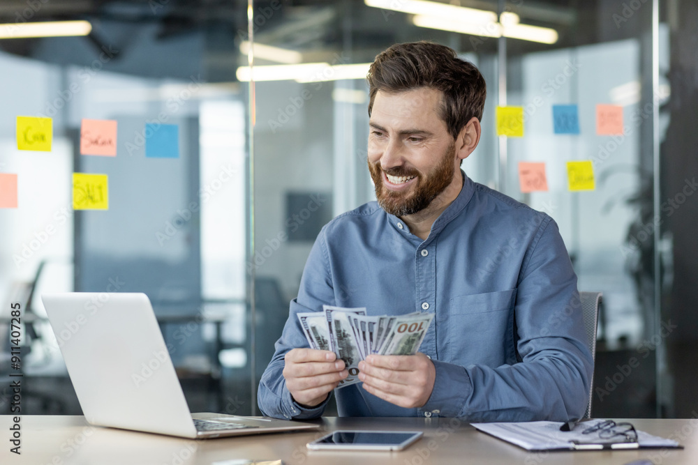 Smiling businessman counting money while working on laptop in office setting Smiling businessman counting money while working on laptop in office setting