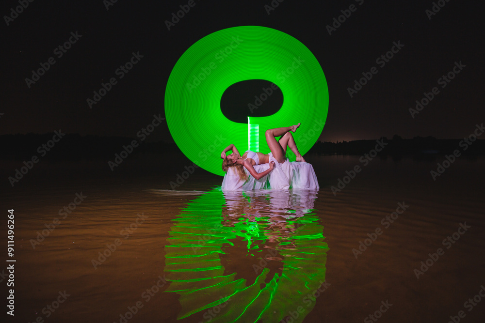 Young slim girl in white swimsuit posing in water at night with abstract green light effect