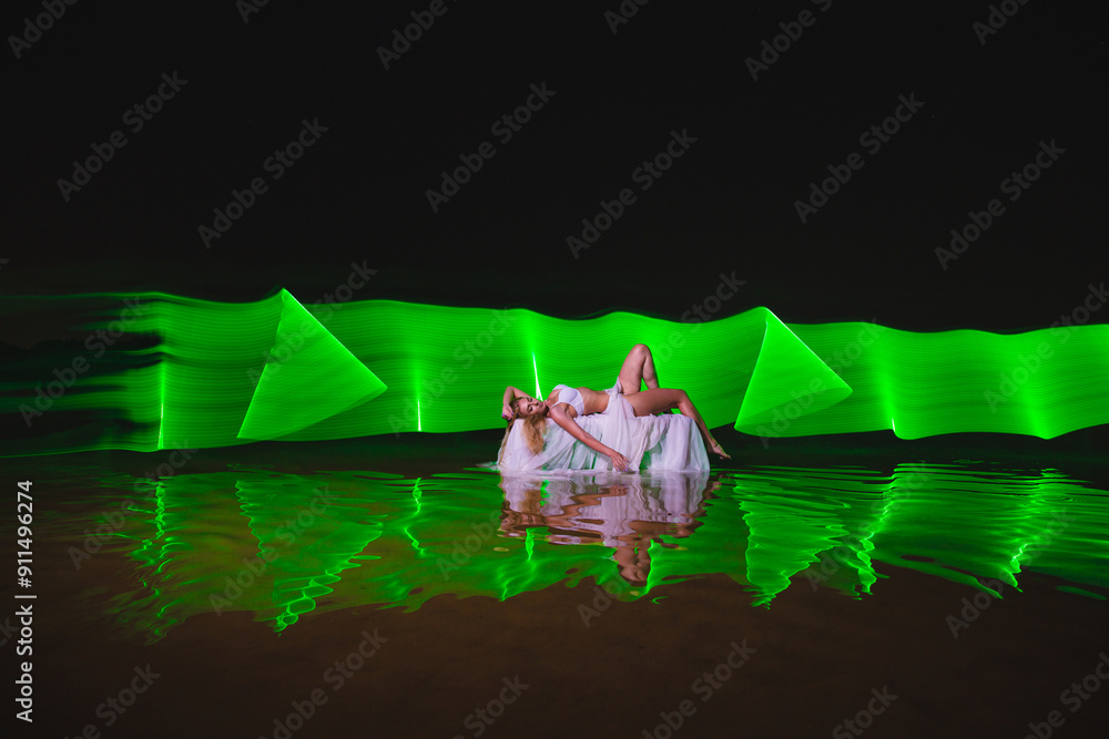 Young slim girl in white swimsuit posing in water at night with abstract green light effect