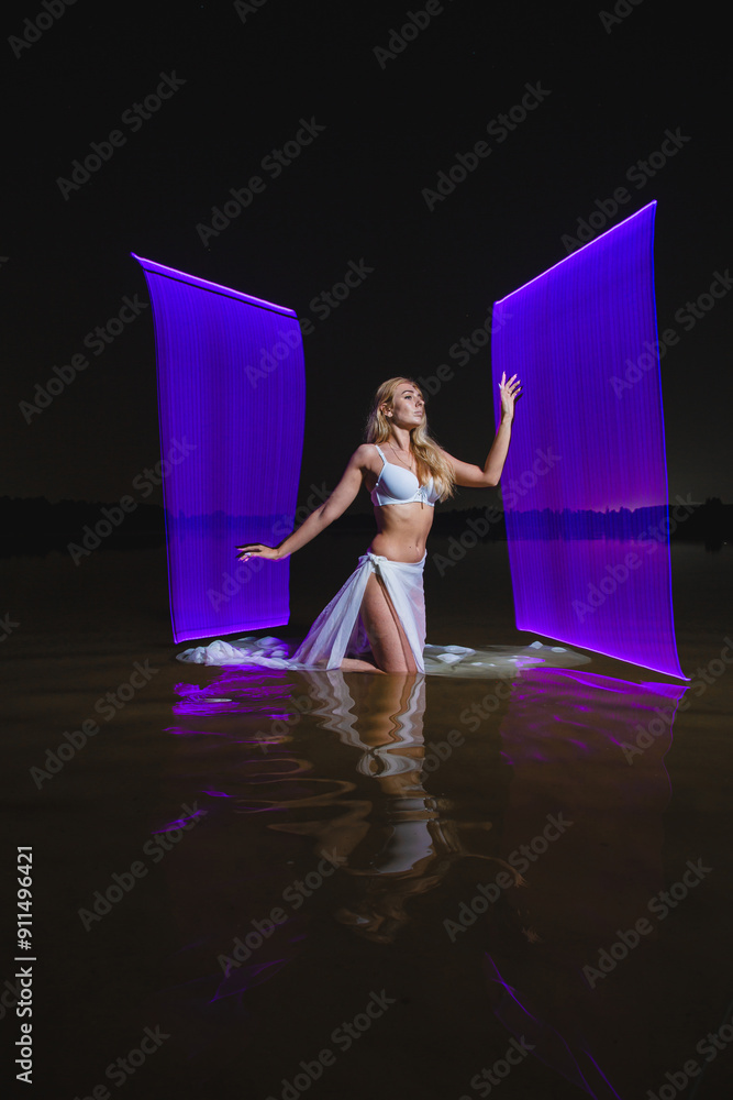 Young slim girl in white swimsuit posing in water at night with abstract purple light effect