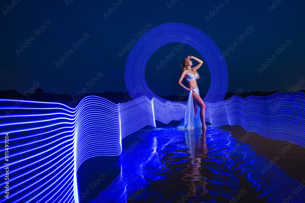 Young slim girl in white swimsuit posing in water at night with abstract blue light effect