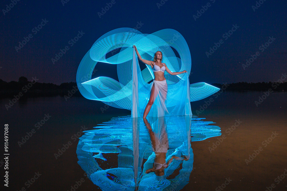 Young slim girl in white swimsuit posing in water at night with abstract blue light effect