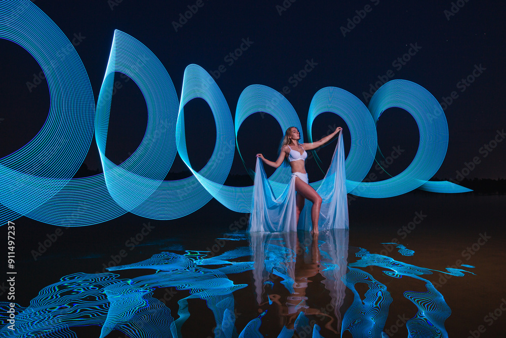 Young slim girl in white swimsuit posing in water at night with abstract blue light effect