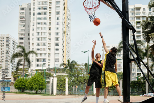 Two young men playing basketball on urban outdoor court surrounded by high-rise buildings. One player is attempting a layup while other is trying to block shot