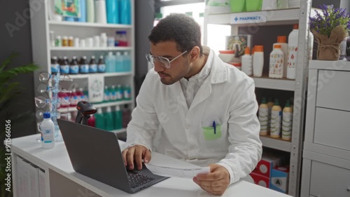 Wallpaper Mural Young man working in a pharmacy shop examining a product while using a laptop and wearing a lab coat and glasses Torontodigital.ca