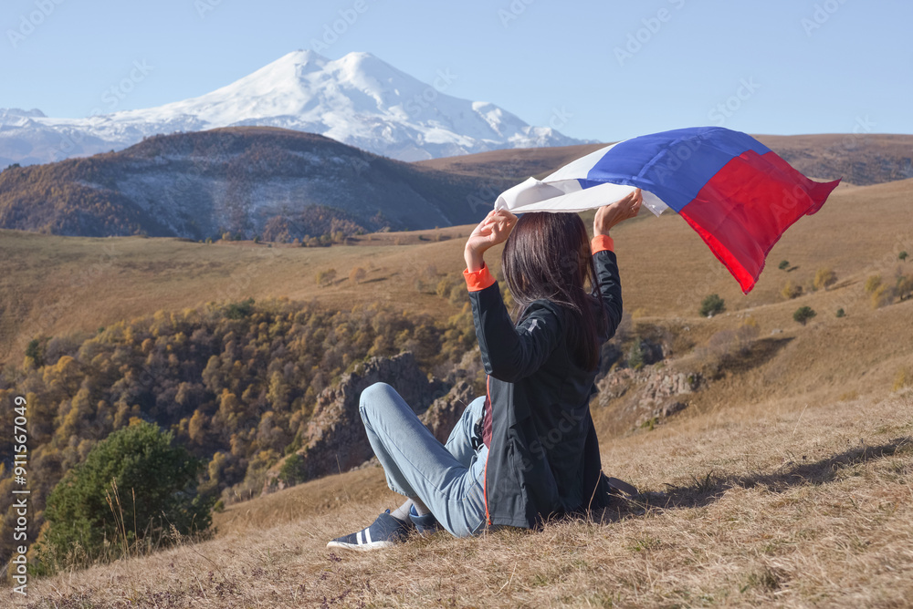 A young brunette woman sits on a stone and looks at the Caucasus Mountains around, a Russian flag flutters in her hands. Tricolor against the backdrop of snow-capped Mount Elbrus