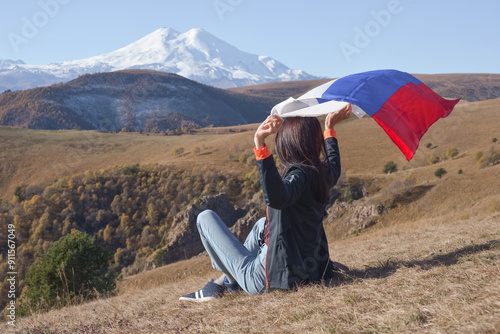 Obraz na płótnie A young brunette woman sits on a stone and looks at the Caucasus Mountains around, a Russian flag flutters in her hands. Tricolor against the backdrop of snow-capped Mount Elbrus w salonie