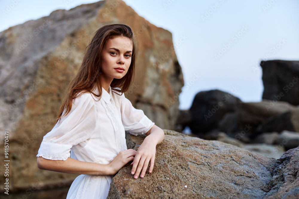 Serene young woman in white dress standing confidently on rocky beach with hands on hips Serene young woman in white dress standing confidently on rocky beach with hands on hips