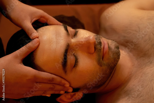 Caucasian man enjoying relaxing anti-stress head massage and pampering facial beauty skin recreation leisure in warm candle lighting ambient salon spa in luxury resort or hotel. Quiescent