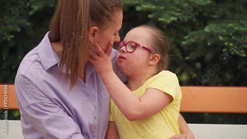 Mother and her young daughter with Down syndrome sit on a colorful bench. Daughter gives her mother a kiss on cheek, reflecting tender and affectionate moments of nurturing child with special needs