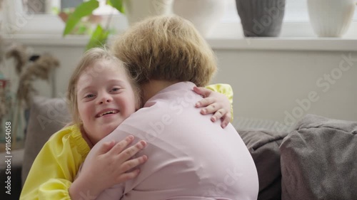Laughing child with Down syndrome hugs her mother in cozy living room. Scene reflects acceptance and the importance of social inclusion within families. Concept: social acceptance, inclusion, support