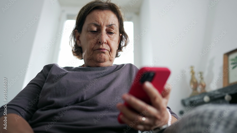 Senior woman holding cellphone device reading content online while seated on couch at living-room, serious expression of 70s person looking at screen Senior woman holding cellphone device reading content online while seated on couch at living-room, serious expression of 70s person looking at screen