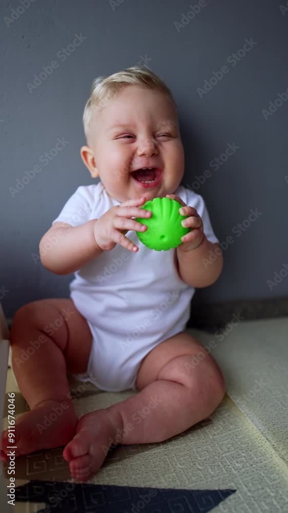 Beautiful blond baby boy wearing white bodysuit sitting on the floor. Kid is laughing adorably playing with a toy. Vertical video. Beautiful blond baby boy wearing white bodysuit sitting on the floor. Kid is laughing adorably playing with a toy. Vertical video.