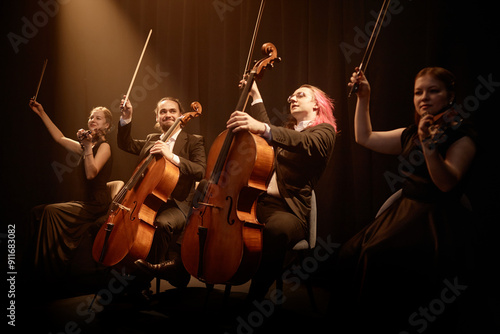 Chamber quartet of string musicians sitting in row on stage with project light raising bows while thanking public at end of performance