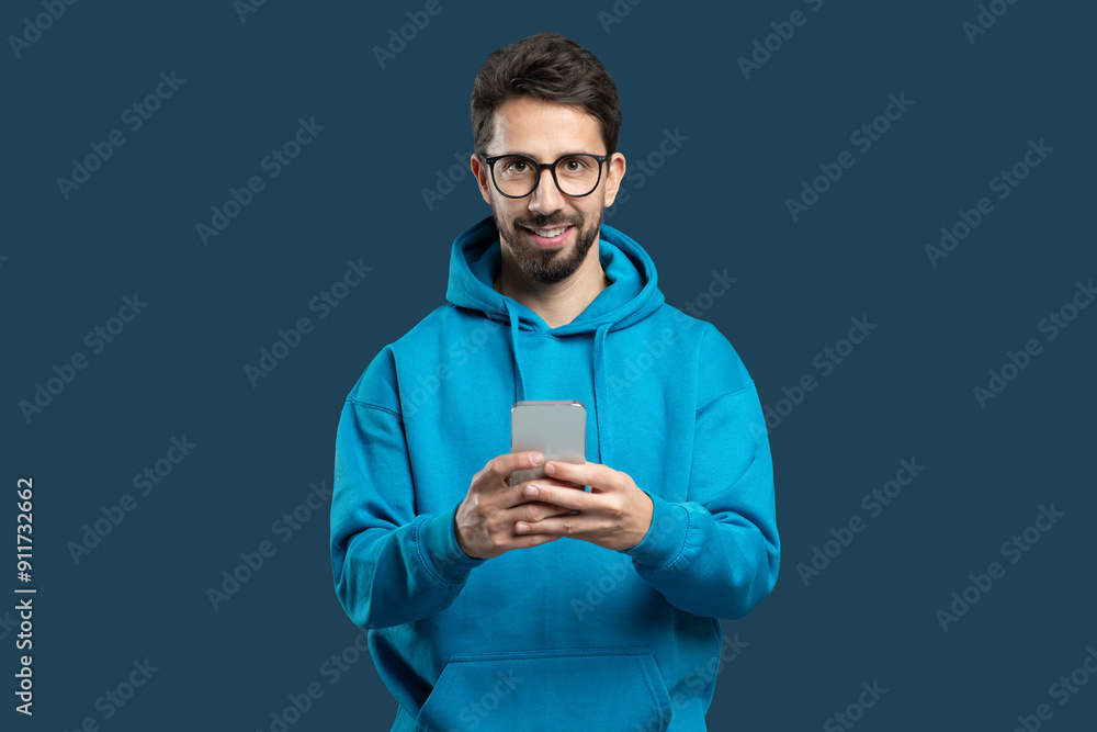 A young man with glasses and a beard stands indoors, wearing a vibrant blue hoodie. He smiles brightly as he interacts with his smartphone, conveying a sense of joy and engagement. A young man with glasses and a beard stands indoors, wearing a vibrant blue hoodie. He smiles brightly as he interacts with his smartphone, conveying a sense of joy and engagement.