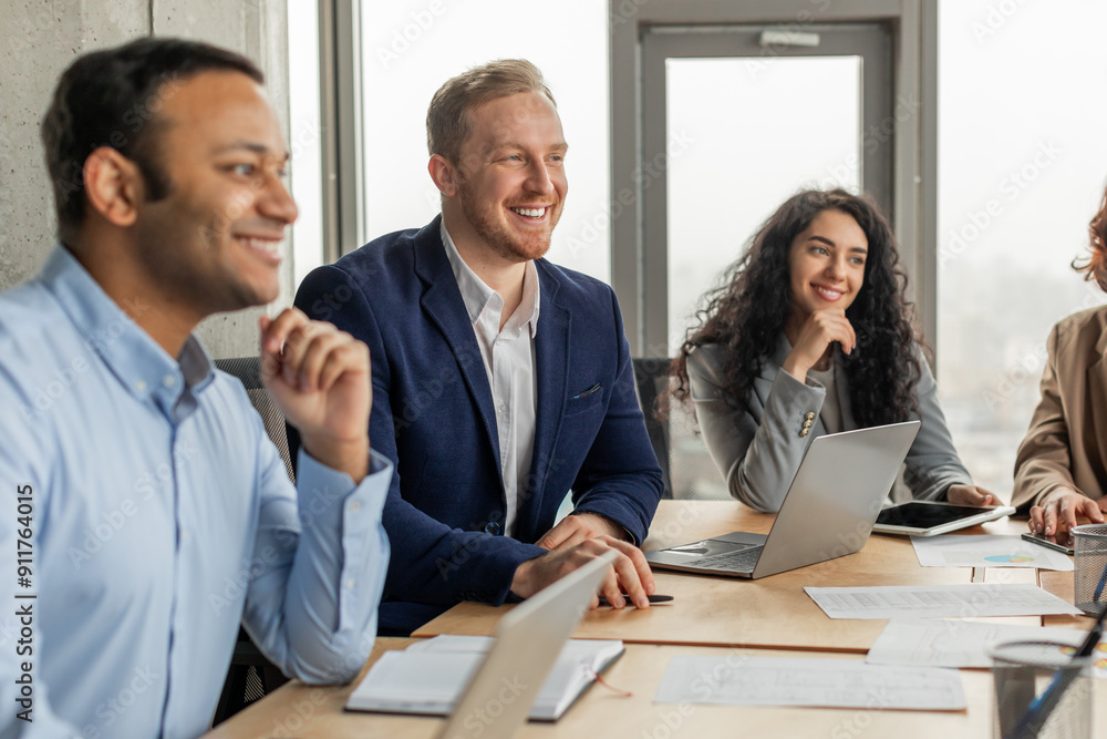 Four smiling colleagues are sitting around a table, engaged in a conversation during a meeting. The man on the left is listening intently, while the man in the middle is speaking Four smiling colleagues are sitting around a table, engaged in a conversation during a meeting. The man on the left is listening intently, while the man in the middle is speaking