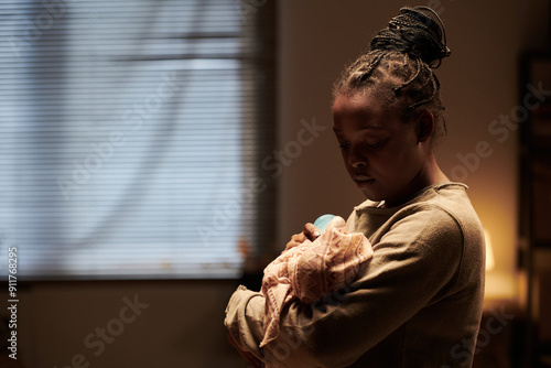 Young African American woman with baby on hands feeding infant with milk while standing in front of camera in home environment