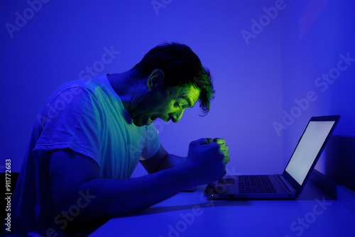 Frustrated Person in White T-Shirt Shouting at Laptop Under Blue Lighting at Night