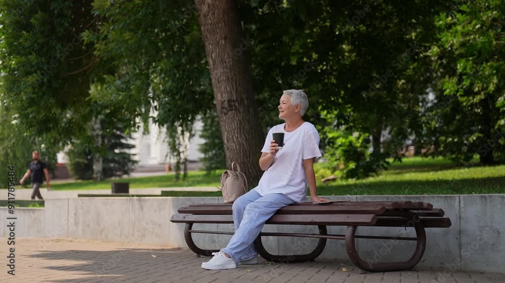 Wide remote shot of serene casual smiling 60s female sitting on bench and drinking coffee outdoor in park on summertime. Happy relaxed older woman drinking takeaway coffee on wooden bench in park. Wide remote shot of serene casual smiling 60s female sitting on bench and drinking coffee outdoor in park on summertime. Happy relaxed older woman drinking takeaway coffee on wooden bench in park.