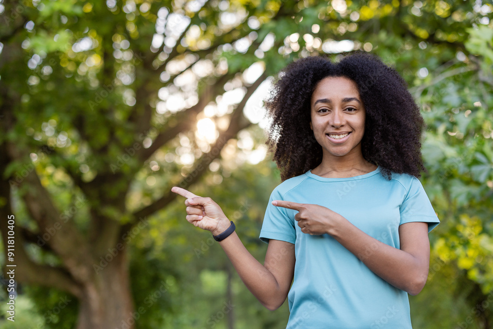 Smiling African American woman in blue shirt pointing sideways in park setting with trees in background. Concept of happiness, outdoors, and positive vibes. Smiling African American woman in blue shirt pointing sideways in park setting with trees in background. Concept of happiness, outdoors, and positive vibes.