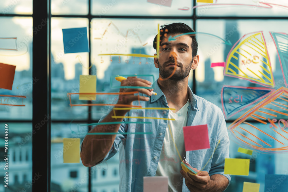 Fototapeta premium Portrait of young handsome businessman writing marketing idea by using colorful marker on glass wall with graph and mind map. Manager sharing idea or financial strategy. Business plan. Tracery.