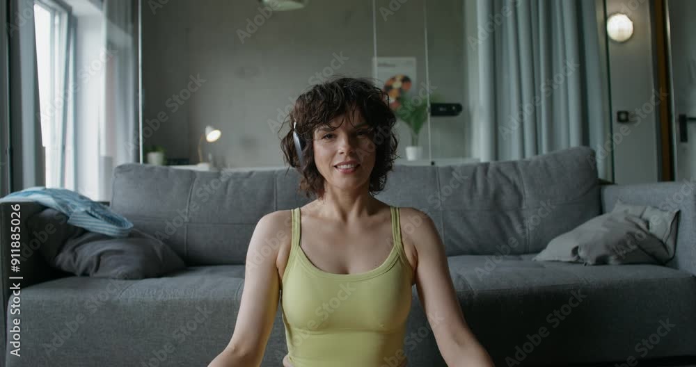 A woman in sports clothes and with headphones on her head is smiling at the camera while sitting in the lotus position at home. Daylight A woman in sports clothes and with headphones on her head is smiling at the camera while sitting in the lotus position at home. Daylight