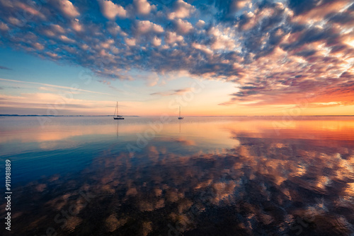 Calm summer seascape of Baltic sea with two yacht on the horizon. Colorful endless clouds in colorful sky reflected in the calm waters of Baltic sea. Fantastic sunrise on Rewa public beach, Poland.