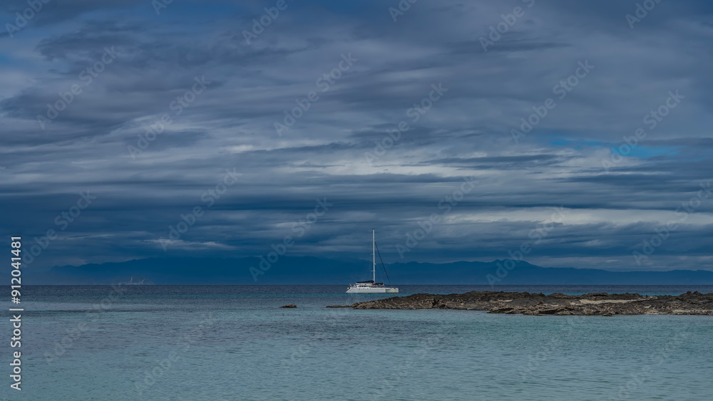 Fototapeta premium A serene seascape. The white yacht is anchored in the calm turquoise ocean. A tall mast against a background of blue sky and clouds. A rocky island. A mountain range in the distance. Madagascar.