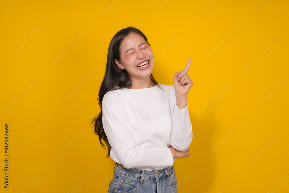 Young woman is laughing and pointing upwards with her index finger against a vibrant yellow background Young woman is laughing and pointing upwards with her index finger against a vibrant yellow background