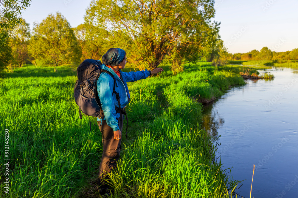 A hiker gestures towards the flowing water, surrounded by vibrant green grass and trees as the morning sun casts a warm light over the serene landscape. A hiker gestures towards the flowing water, surrounded by vibrant green grass and trees as the morning sun casts a warm light over the serene landscape.