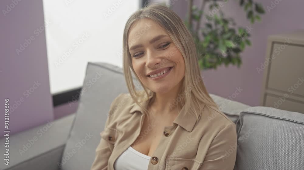 Young blonde woman sitting on sofa, smiling and looking to the side, thinking away at home Young blonde woman sitting on sofa, smiling and looking to the side, thinking away at home
