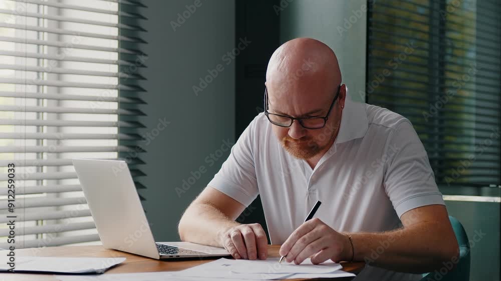 Busy adult man works sitting at the desk at workplace in home office. A serious financier checks documents, does paper work. Businessman working on startup or project. Busy adult man works sitting at the desk at workplace in home office. A serious financier checks documents, does paper work. Businessman working on startup or project.