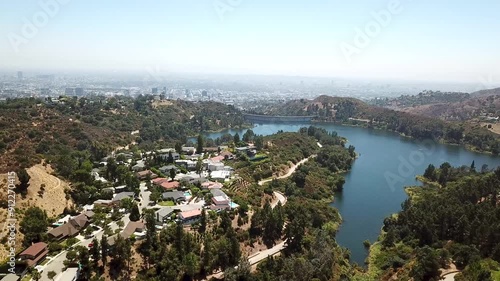 Aerial establishing shot of luxury neighborhood with mansions and Hollywood reservoir during sunny day. Los Angeles Cityscape in background. Drone wide shot.