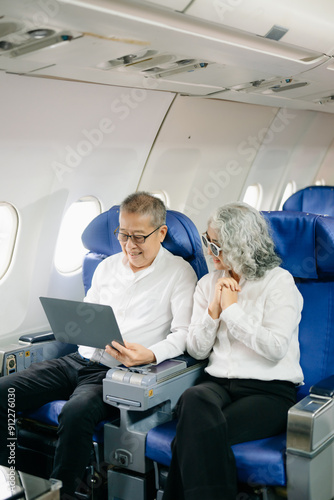 Smiling senior couple of Asian tourists take photos of each other while sitting on a plane. with digital tablet, laptop and smartphone. Travel in style,