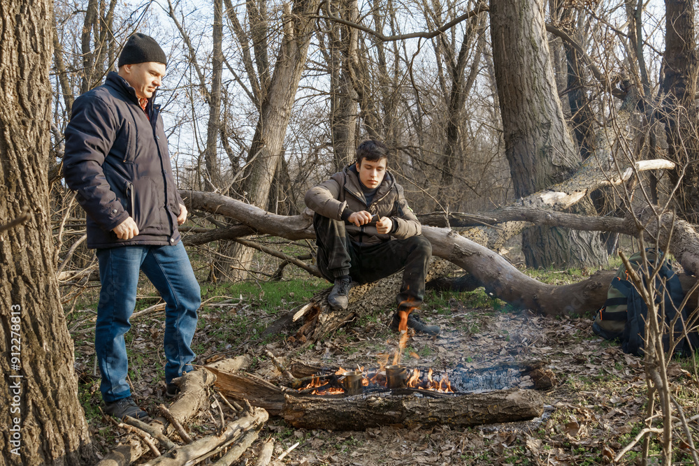 Two men make a campfire for cooking in the forest, father and son sitting on a log while hiking and outdoor activities, early spring landscape Two men make a campfire for cooking in the forest, father and son sitting on a log while hiking and outdoor activities, early spring landscape