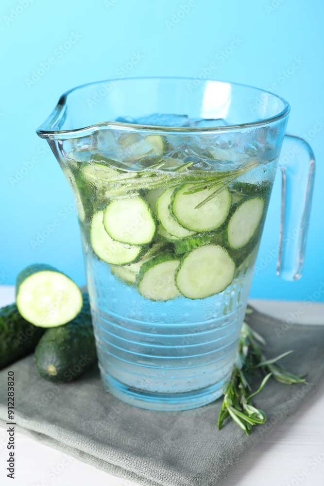 Refreshing cucumber water with rosemary in jug and vegetables on white table against light blue background Refreshing cucumber water with rosemary in jug and vegetables on white table against light blue background